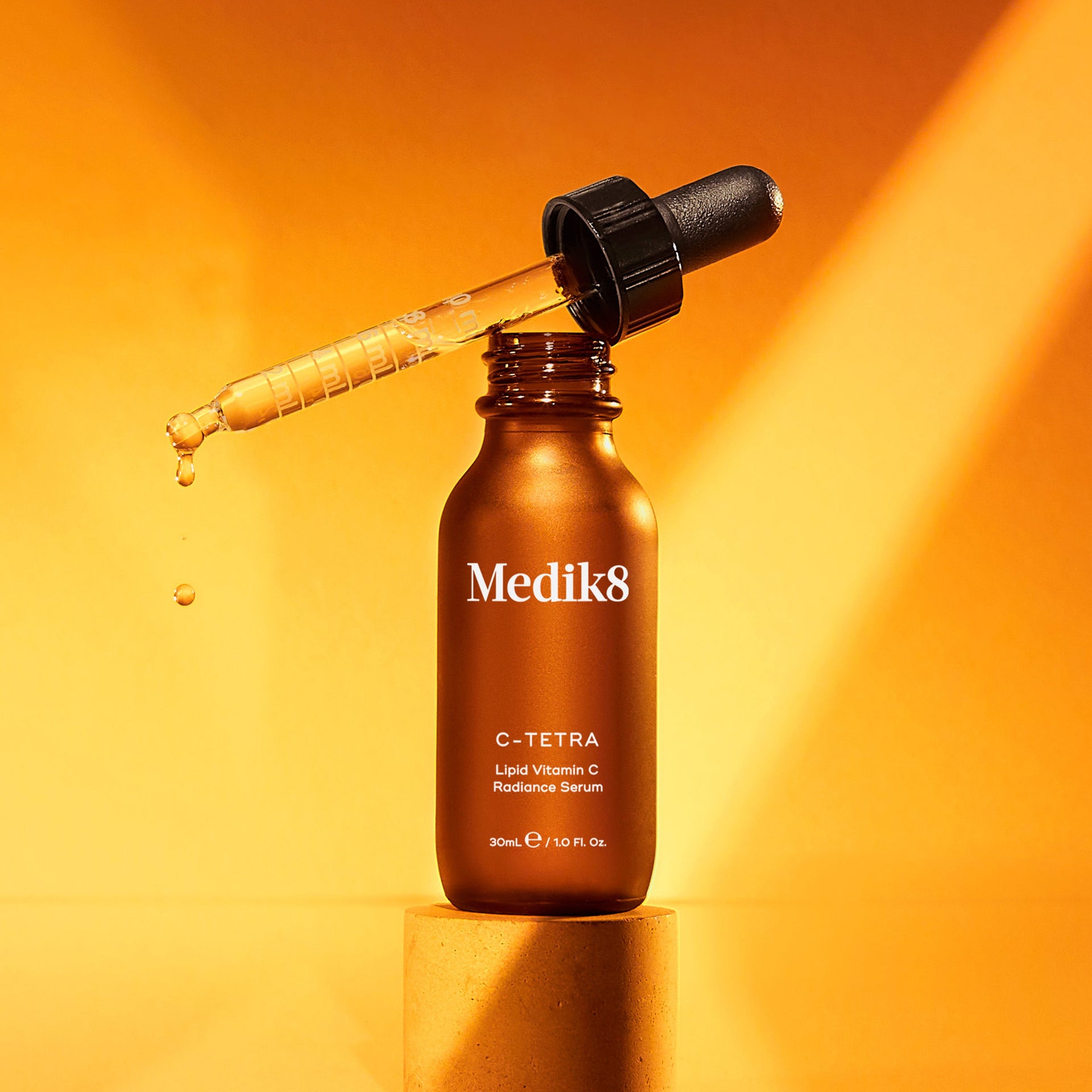 En brun glasflaska Medik8 C-Tetra, ett serum med vitamin C, står på en cylinderformad plattform. Pipetten ovanför flaskan droppar försiktigt produkten. Bakgrunden är en varm orange färg med ljuseffekter.