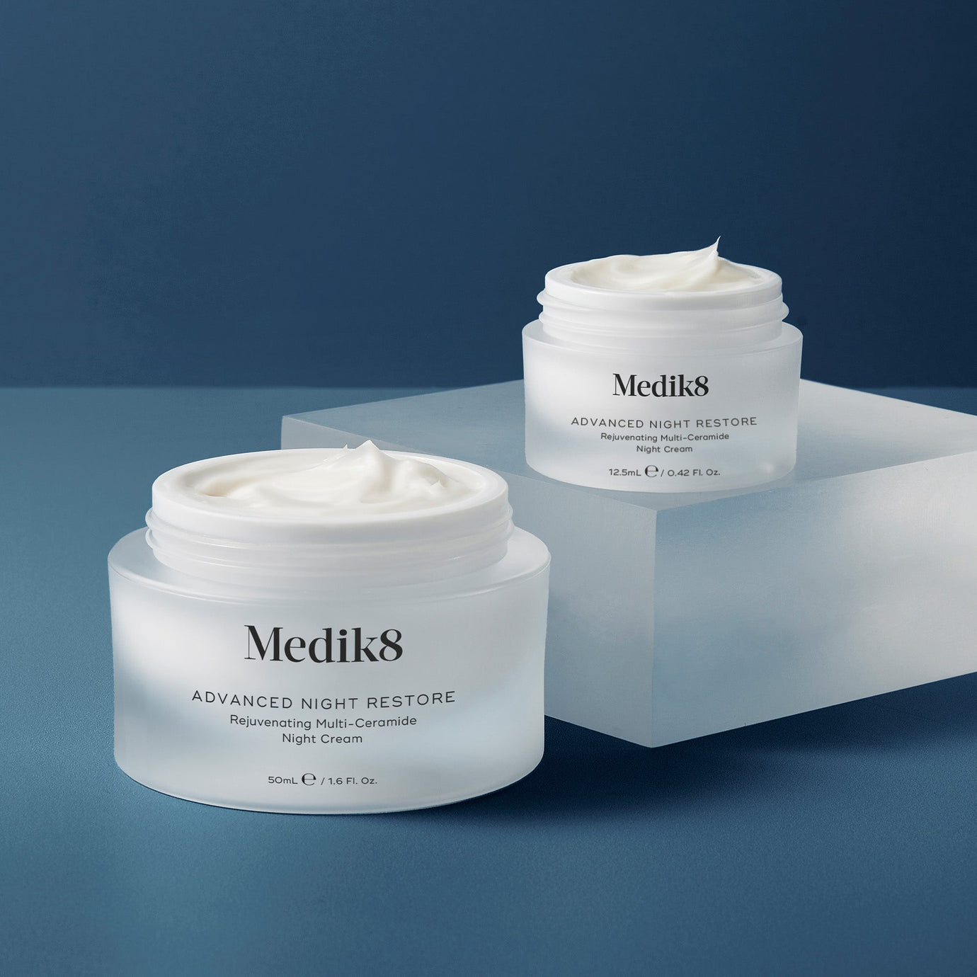 Två burkar av Medik8 Advanced Night Restore nattkräm i olika storlekar på en blå bakgrund. Två burkar av Medik8 Advanced Night Restore nattkräm i olika storlekar på en blå bakgrund.