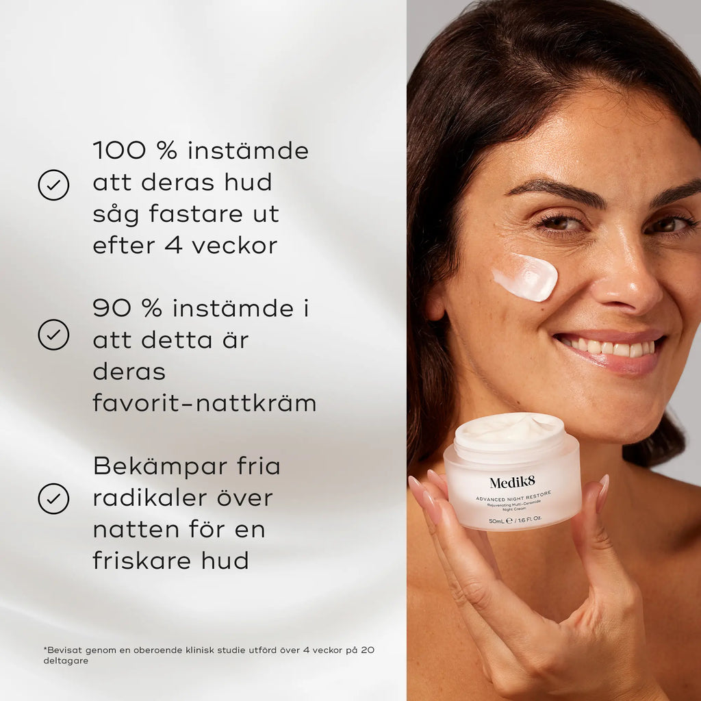Leende kvinna applicerar Medik8 Advanced Night Restore nattkräm på kinden medan hon håller i en burk med produkten. Bakgrunden har en vit, silkeslen textur med text om produktens hudförbättrande egenskaper.