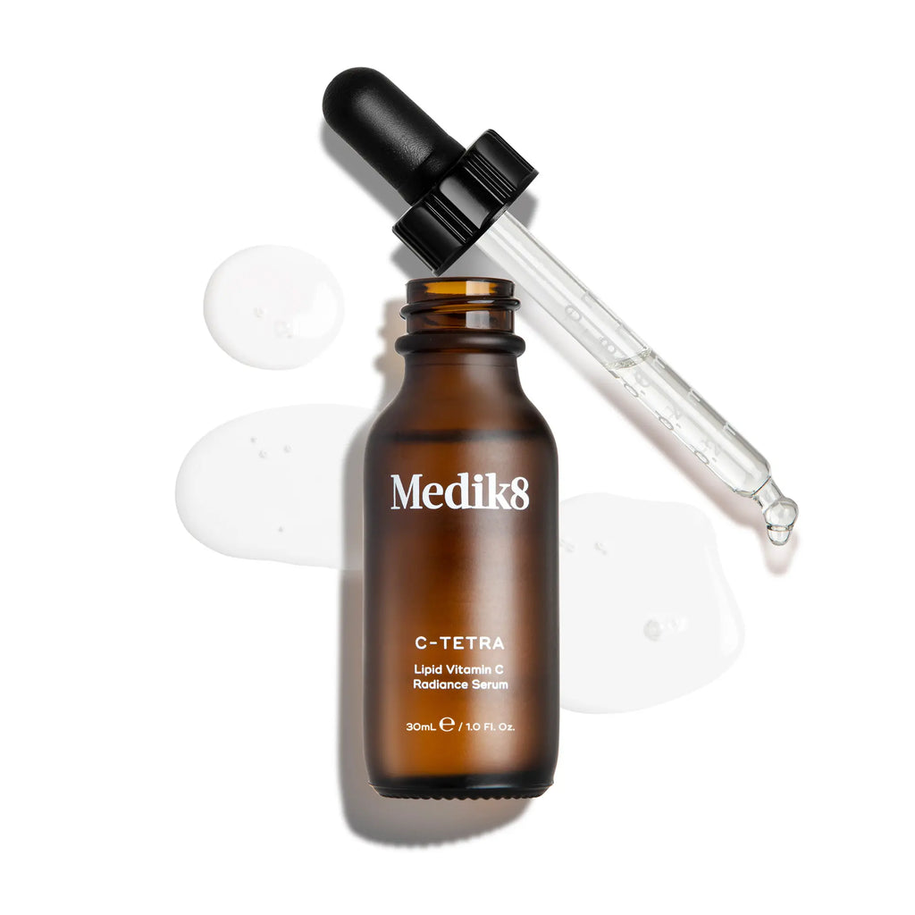 Medik8 C-Tetra Vitamin C-serum i brun glasflaska med pipett, omgiven av genomskinliga droppar serum.