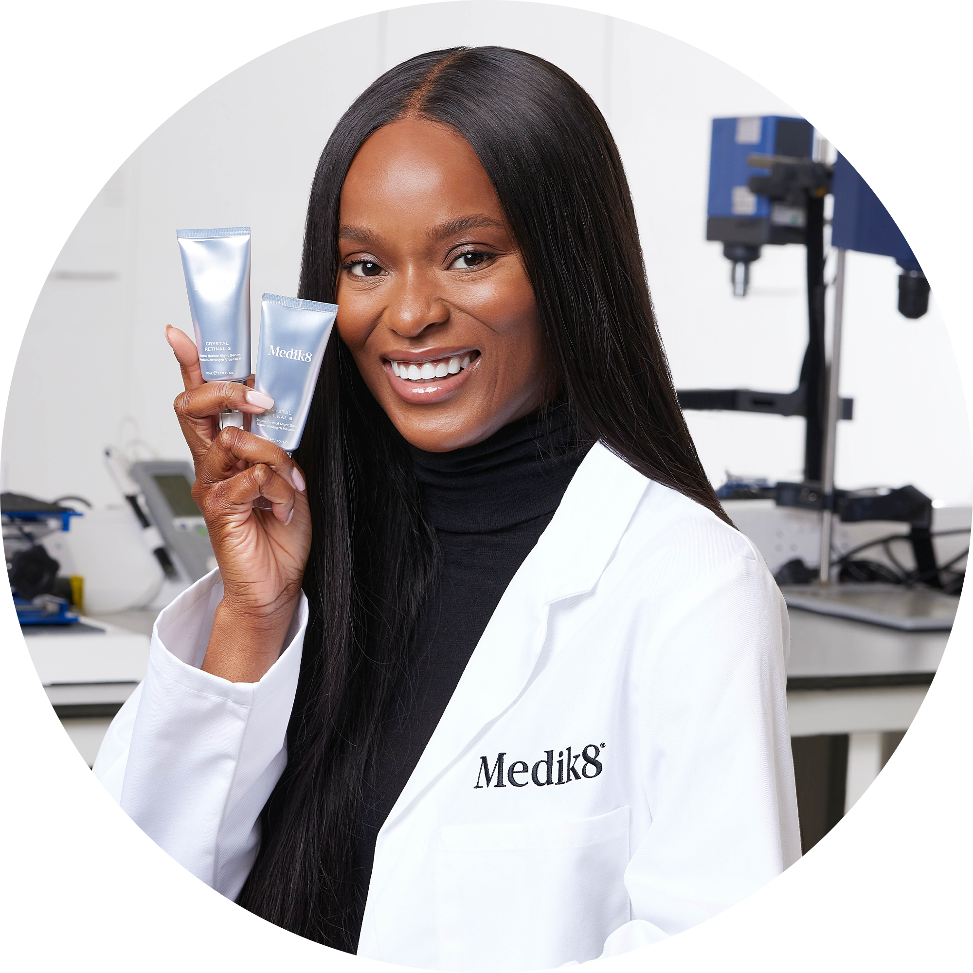 Dermatolog i vit laboratorierock från Medik8 håller två silverfärgade tuber med hudvårdsprodukter i ett laboratorium.