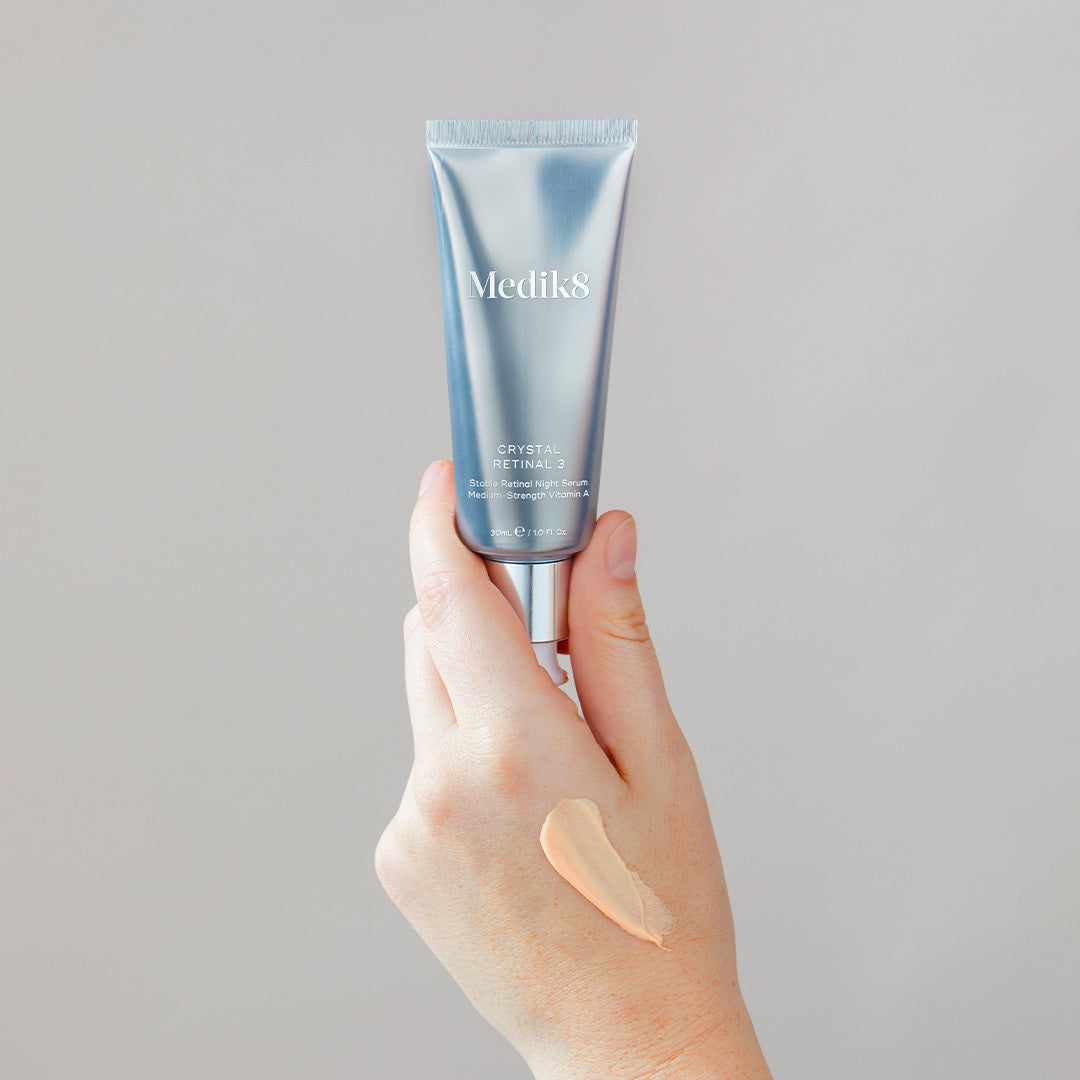 En hand håller en tub Medik8 Crystal Retinal 3 med en klick av produkten applicerad på handen. Bakgrunden är neutral.
