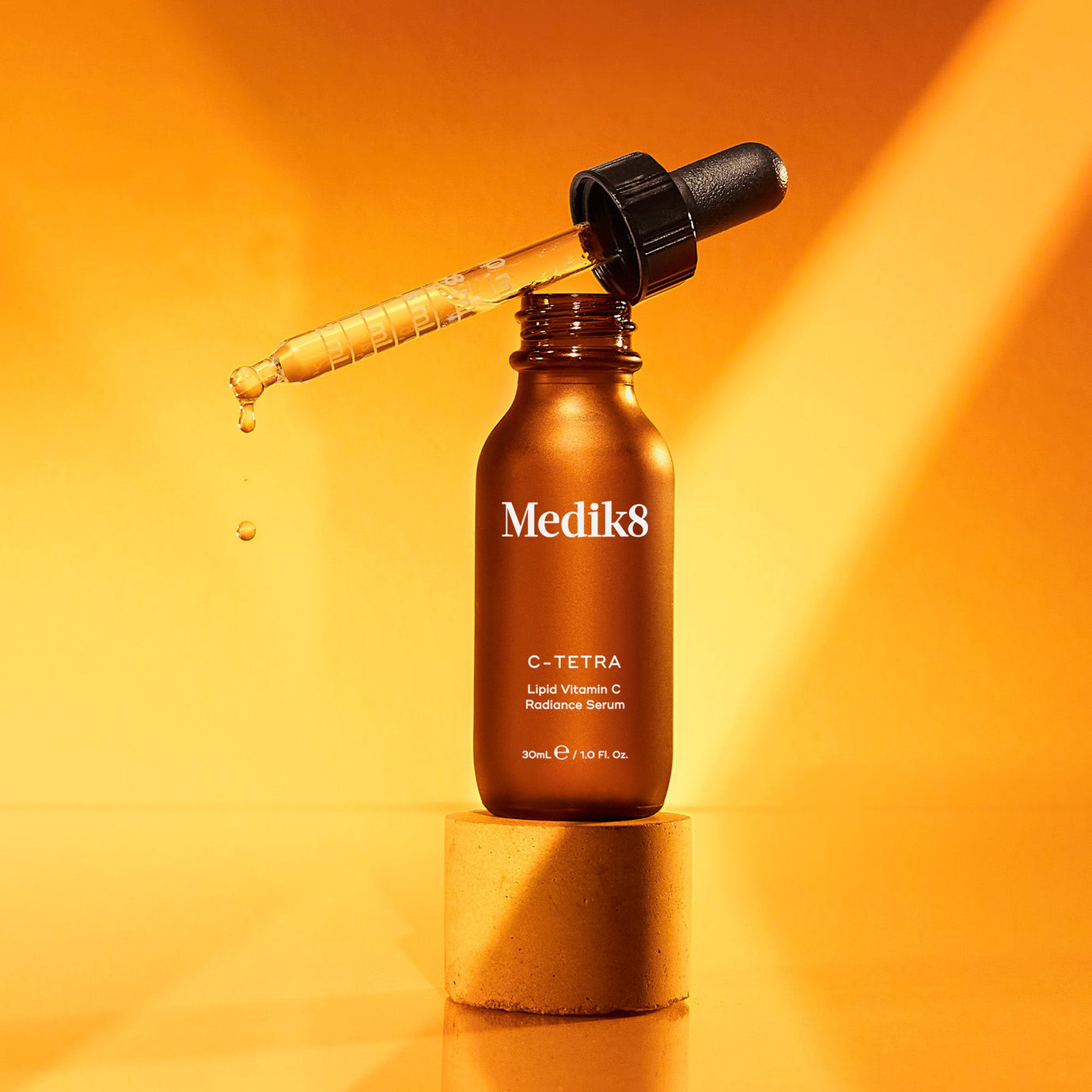 Flaska med Medik8 C-Tetra Vitamin C-serum på en orange bakgrund, med en pipett som droppar serum.